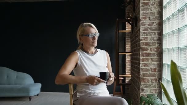 Όμορφη γυναίκα πίνει καφέ κάθεστε δίπλα στο παράθυρο κοιτάζοντας έξω με ευτυχισμένη έκφραση. Γυναίκα ώριμη επιχείρηση απολαμβάνοντας καφέ κάθεται στο γραφείο κατά τη διάρκεια της διάλειμμα για καφέ — Αρχείο Βίντεο