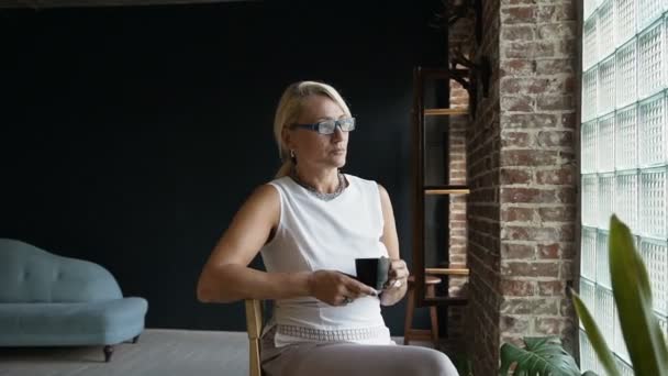 Härlig äldre kvinna dricker kaffe sitter nära fönstret tittar utanför med glad uttryck. Mogen business kvinna njuter kaffe sitter på kontor kafferast — Stockvideo