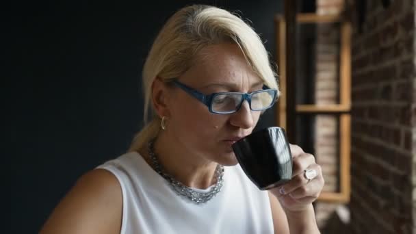 Retrato de mujer encantadora de mediana edad en gafas es beber café sentado cerca de la ventana mirando hacia el exterior con expresión feliz. Mujer de negocios disfrutando de café sentado en la oficina durante el descanso del café — Vídeos de Stock