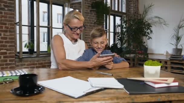 Donna caucasica di mezza età seduta a tavola nell'accogliente stanza dell'ufficio e che racconta qualcosa al suo grazioso nipote adolescente mentre dimostra qualcosa sullo schermo del tablet del computer — Video Stock