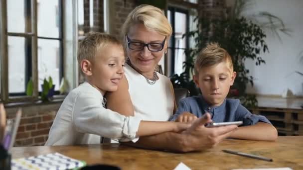 Портрет прекрасной бабушки с внуками, использующих планшет для видеочата, сидя на столе дома в офисной комнате — стоковое видео