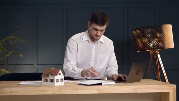 Un agente de bienes raíces masculino serio haciendo alguna nota en el cuaderno de la computadora portátil . — Vídeos de Stock