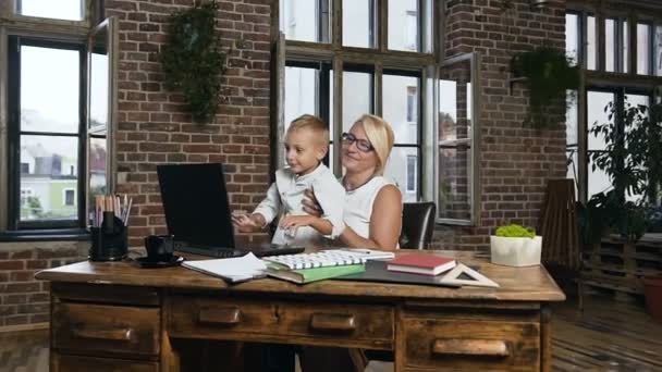 Красивая деловая женщина средних лет сидит за деревянным столом и наблюдает за своим милым внуком, играющим на ноутбуке в ее офисе — стоковое видео