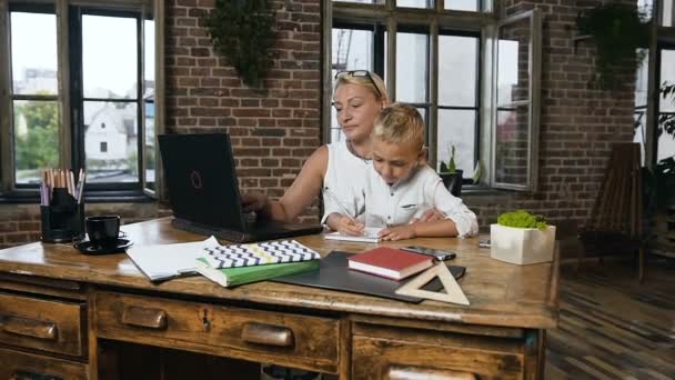 Mooie middelbare leeftijd zakenvrouw die op laptop werkt wanneer haar kleine mooie kleinzoon iets in een notitieblok op kantoor schrijft. Dolly geschoten, binnenshuis — Stockvideo