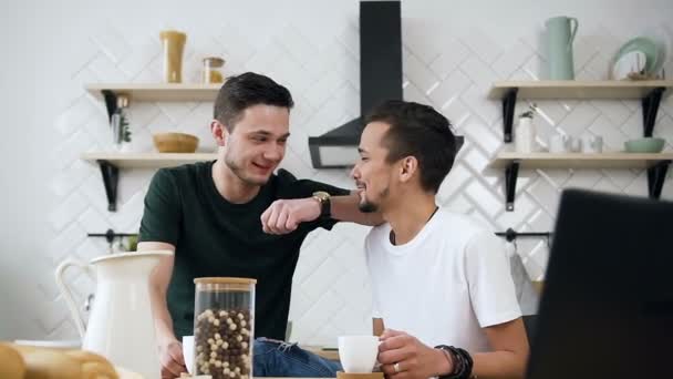 Young male couple are sitting at the kitchen table talking something fun while drinking coffee in the morning at kitchen. Comfort and relaxation — Stock Video