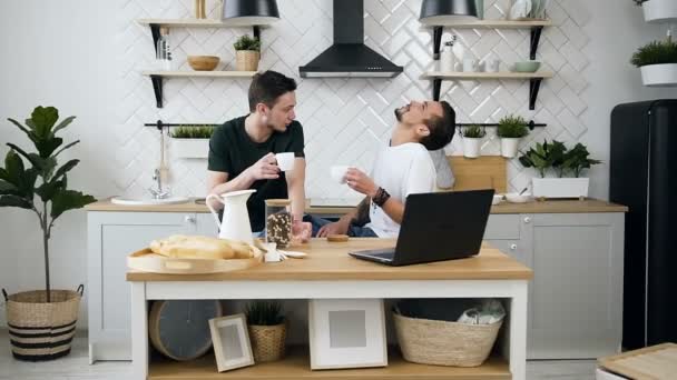 Alegre casal gay de homens bonitos estão sentados atrás da mesa da cozinha algo falando para durante a bebida de café na cozinha pela manhã. Colegas de quarto bebendo café juntos pela manhã em — Vídeo de Stock