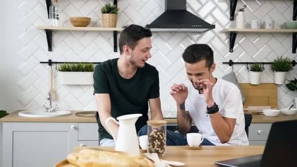 Quelques beaux hommes gays sont assis derrière la table de cuisine quelque chose qui parle pendant la consommation de café à la cuisine le matin. Colocataires buvant du café ensemble le matin à la cuisine — Video