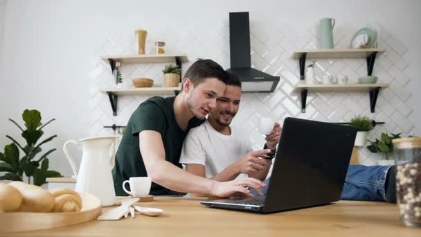 Молодая пара геев на кухне обнимаются и завтракают за обеденным столом, пользуясь интернетом на ноутбуке. Они забавно улыбаются и разговаривают. В помещении. Светлый кухонный фон — стоковое видео