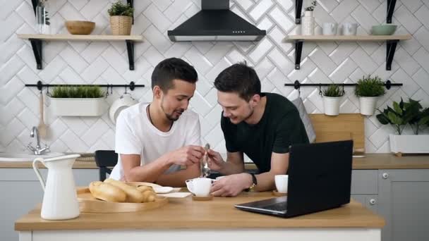 Portret kaukaski para gejów siedzą za stołem kuchennym i o śniadanie rano w kuchni. Przystojny młody facet jest karmienie swojego partnera siedzi przy stole w kuchni rano — Wideo stockowe