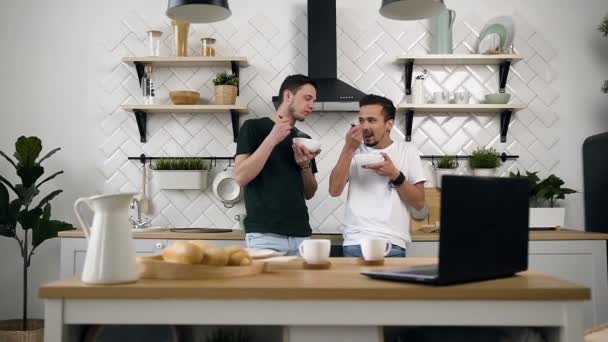 Щасливі молоді гей-партнери, що стоять біля кухонного столу, щось говорять і снідають вдома вранці на кухні — стокове відео