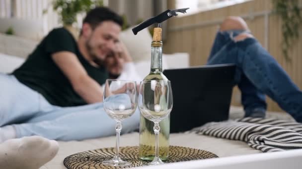 Bottle wine with two glass standing on lovely gay couple bed. Attractive gay couple using laptop together with funny emotion laying in the bed at bedroom. Gay men spending time together — Stock Video