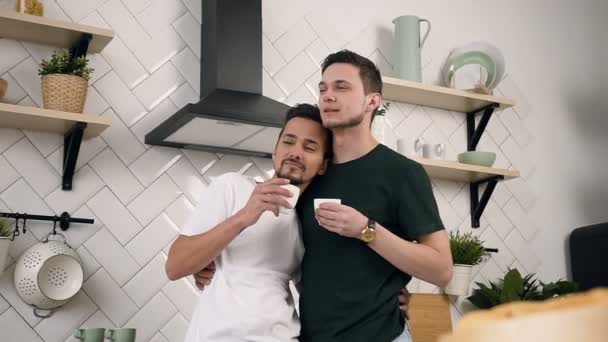 Portrait of young male gay couple are hugging and drinking morning coffee while standing at the kitchen at home — Stock Video