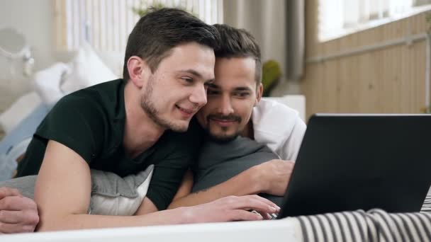 Casal jovem homossexual assistindo filme engraçado no computador portátil deitado na cama. Caucasiano divertindo casal gay assistindo na tela do laptop enquanto descansa na sala de estar deitado no sofá — Vídeo de Stock