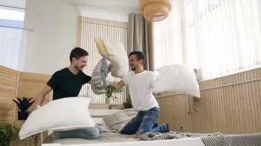 Genç gay çift yatak funing gülüyor, flört ve mücadele yastık ışık evinde oturan. Yastık sahip mutlu gay çift Çift Kişilik Yatak üzerinde gülen ve having fun, mücadele. İnsanlar, romantik