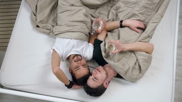 Mutlu genç homoseksüel erkekler çift beyaz şarap içme ve geleceği hakkında hayal ederken konuşmak ışık yatak odası yatakta yatan görüntüsünü el Üstten Görünüm — Stok video