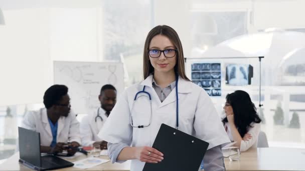 Porträt einer attraktiven jungen Frau, die in die Kamera blickt und sich die Hände kreuzt, während das Team im Krankenhaus im Hintergrund arbeitet. — Stockvideo