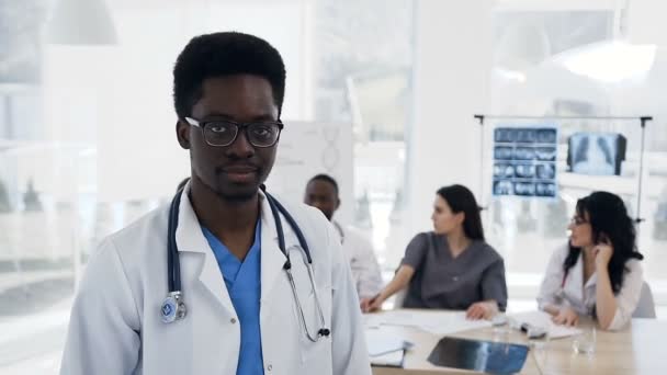 Çekici Afrika erkek doktor hastane arka planda cowoker ekibi ile kameraya bakıp. — Stok video