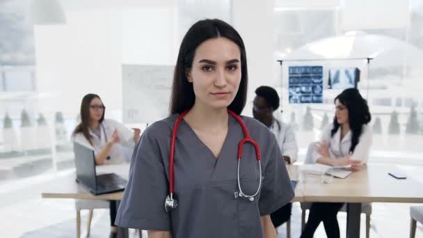 Женщина-врач смотрит в камеру и скрещивает руки, пока команда сотрудников работает на заднем плане в больнице . — стоковое видео
