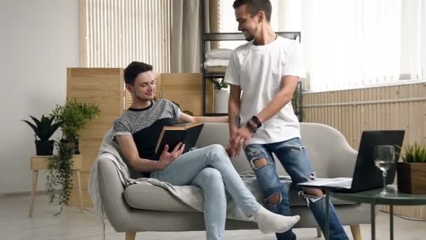 Gelukkige homo paar mannen zijn het lezen van boek op de Bank samen. Jonge knappe homo man zittend op de Bank lezen boek tijdens zijn homo partner benaderd voor hem ging zitten volgende en horloges wat hij aan het lezen was — Stockvideo