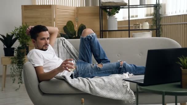 Молодий привабливий чоловік лежить на дивані, розслабляється і п'є ігристе біле вино за переглядом фільму в ноутбуці у вітальні — стокове відео
