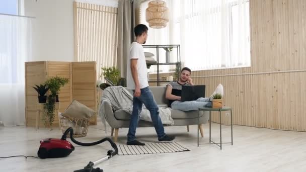 Homoseksualnych par, gejów, same małżeństwa homoseksualne między kaukaski mężczyzn. Dwaj młodzi mężczyźni siedzą na kanapie za pomocą smartfona i laptop po oczyszczeniu w apartamencie — Wideo stockowe