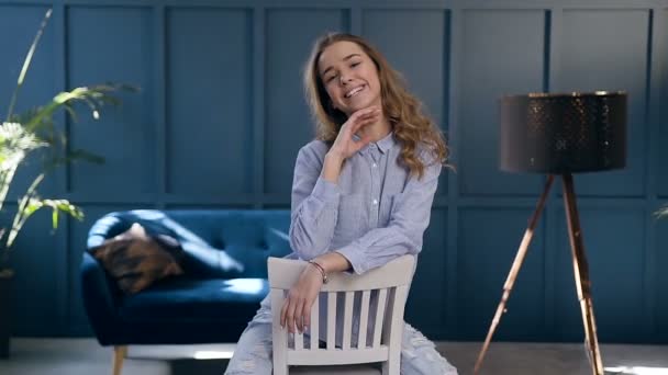 Mulher de cabelo loira feliz sorrindo para a câmera enquanto sentada na cadeira no quarto em casa . — Vídeo de Stock