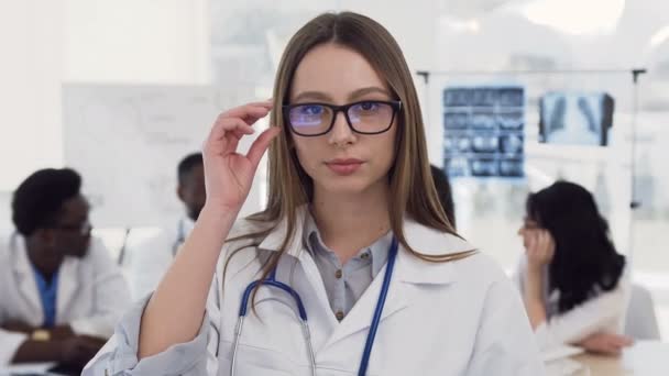 Портрет уверенной женщины-врача в очках и белом халате со стетоскопом на шее, улыбающейся в камеру на фоне коллег в больнице. Врач, здравоохранение, любовь к медицине — стоковое видео