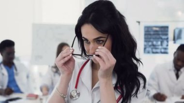 Gözlük güzel bir genç kadın doktor portre kamera bakıyor, onun meslektaşları konferans klinikte arka planda masanın arkasında oturan sohbet ederken gülümsüyor