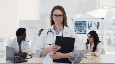 Stetoskop ile beyaz ceket genç kadın doktor klinikte meslektaşları arka planda kamera bakarak gülümsüyor. Doktorlar sağlık kavramı, tıp aşkı