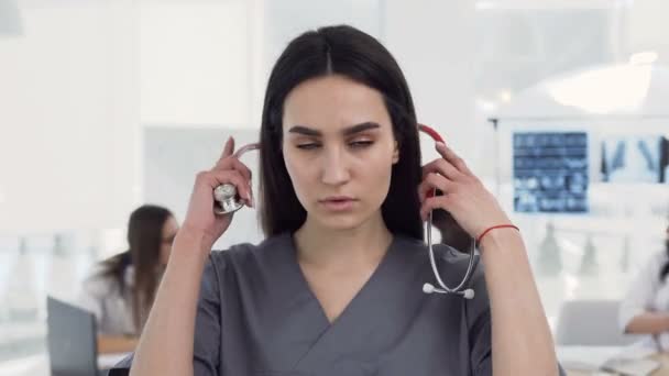 Médica branca com estetoscópio no pescoço em roupas médicas olhando para a câmera e cruzando as mãos enquanto a equipe de funcionários trabalha em segundo plano no hospital — Vídeo de Stock