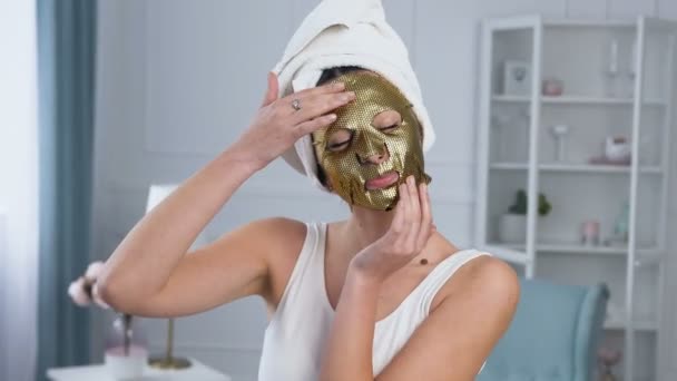 Retrato de mulher sorridente bonito fixando máscara de tecido dourado cosmético rejuvenescedor no rosto . — Vídeo de Stock