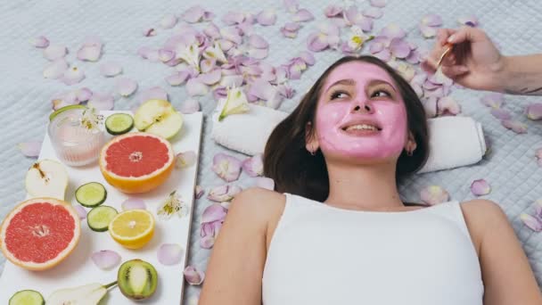 Jovem feliz relaxando no centro de bem-estar, enquanto esteticista aplicando máscara rosa . — Vídeo de Stock
