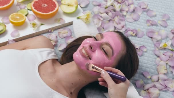 Portret van schattige Kaukasische vrouw ontspannen in Wellness-centrum, terwijl schoonheidsspecialiste toepassing van roze masker met make up Brush. — Stockvideo