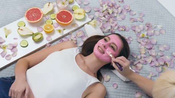 Bonita jovem relaxante no centro de bem-estar, enquanto esteticista aplicando máscara rosa usando maquiagem escova . — Vídeo de Stock