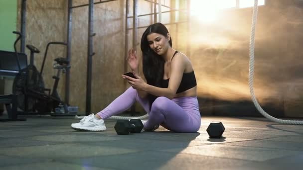 Leende sport kvinna vilar efter träning och använda smarta telefonen för att chatta i gymmet. — Stockvideo