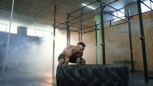 Hombre caucásico fuerte volteando neumático grande durante el entrenamiento en el gimnasio . — Vídeos de Stock