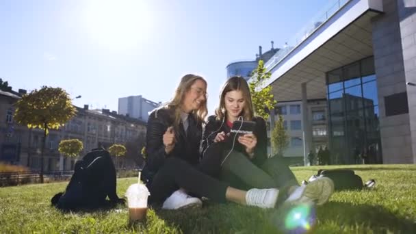 Dos alegre chica universitaria caucásica hablando con auriculares charlando juntos compartiendo cosas divertidas en el dispositivo de la tableta al aire libre en el centro de la ciudad — Vídeos de Stock