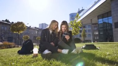 Tablet bilgisayar kullanan sosyal beyaz kız arkadaşlar güneşli havalarda parkta açık havada oturan tabletteki eğlenceli resimlere gülüyorlar