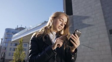 Genç ve güzel bir kadın şehirde yürüyor ve akıllı telefon kullanıyor. Açık havada uygulamayı kullanarak akıllı telefondan mesaj atan gülümseyen güzel kızın portresi. Yaşam tarzı, kentsel