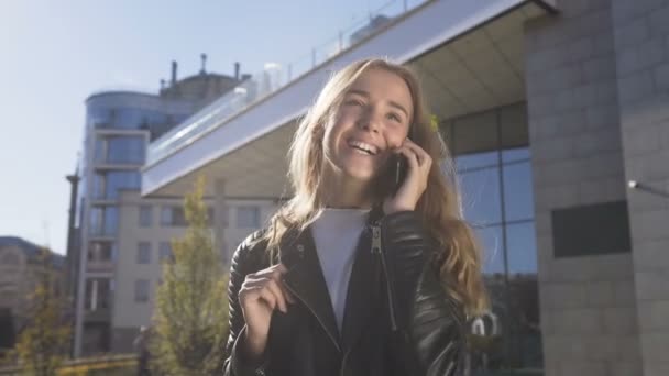 Retrato de una hermosa joven está caminando por la calle de la ciudad mientras habla con su amigo en el teléfono inteligente. Chica rubia bonita hablando en el teléfono inteligente y sonriendo mientras camina al aire libre — Vídeo de stock