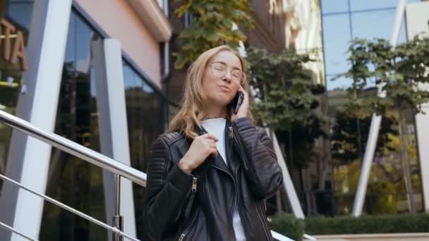 Jeune étudiante séduisante dans des lunettes élégantes avec un long cheveu parlant au téléphone tout en marchant sur le fond de la rue urbaine. En plein air, mode de vie — Video