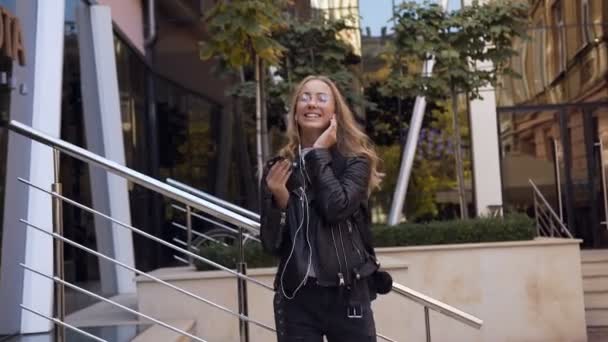 Menina muito atraente em óculos está ouvindo música do telefone em fones de ouvido enquanto caminha na rua urbana. Estilo de vida — Vídeo de Stock