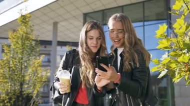 İki neşeli çekici Kafkas kız en iyi arkadaş modern binanın yakınındaki parkta yürüyüş çok gülüyor ve akıllı telefon ekranında bir şey izliyor. Açık havada. Dolly atış