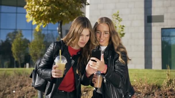 Retrato de las dos elegantes alumnas mejores amigas bebiendo jugo fresco, leyendo algo en el smartphone y riendo. Al aire libre. Dolly disparó — Vídeos de Stock