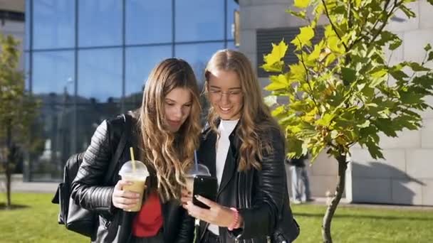 Two young smiling women browsing internet on mobile phone while walking in a park of the city near modern office centre. Outdoors — Stock Video