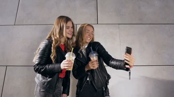 Dos atractivas sonrisas chicas caucásicas tomando fotos selfie en el teléfono inteligente cerca de la pared gris. Al aire libre. Personas, amigos y concepto de estilo de vida — Vídeo de stock