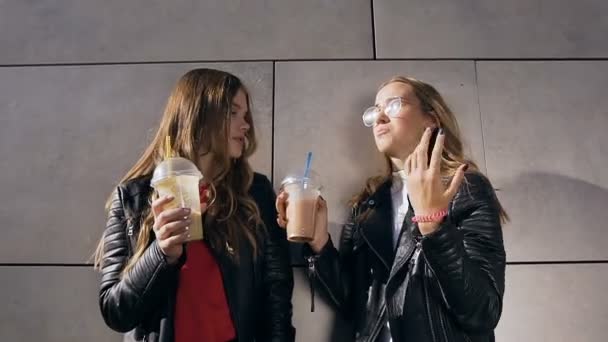 Dwie piękne szczęśliwe dziewczyny rozmawiają i piją koktajle stojąc w pobliżu szarej ściany obok nowoczesnego budynku. Zewnątrz — Wideo stockowe