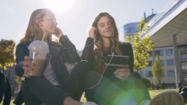Tablet bilgisayar kullanan sosyal beyaz kız arkadaşlar güneşli havalarda parkta açık havada oturan tabletteki eğlenceli resimlere gülüyorlar
