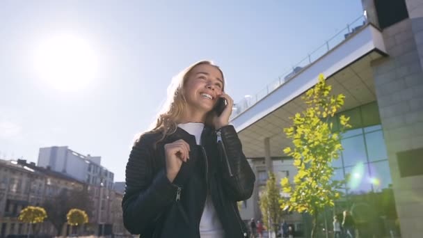Retrato de una bonita chica rubia hablando en smartphone y sonriendo mientras camina por la calle y disfruta del estilo de vida urbano de la ciudad. Al aire libre — Vídeo de stock