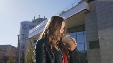 Çekici öğrenci kız şehir parkında yürürken bir süt kokteyli içme ve onun akıllı telefon kulaklık uygulaması üzerinden müzik dinlerken.